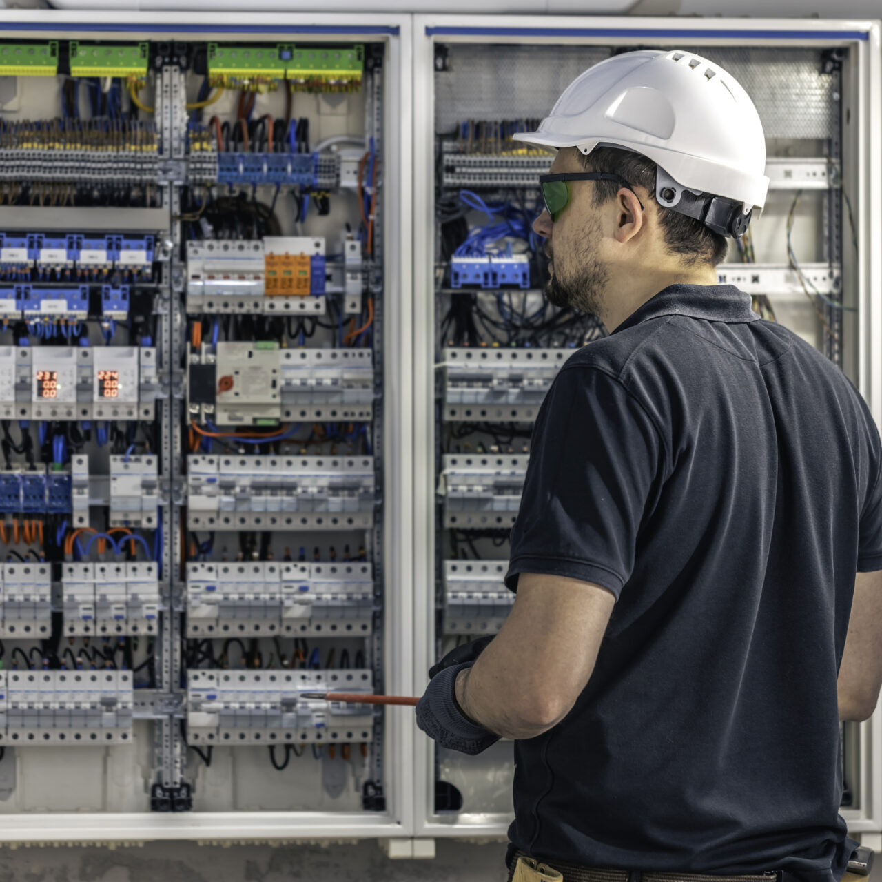 A male electrician works in a switchboard using an electrical connection cable, connecting equipment with tools. View from the back.
